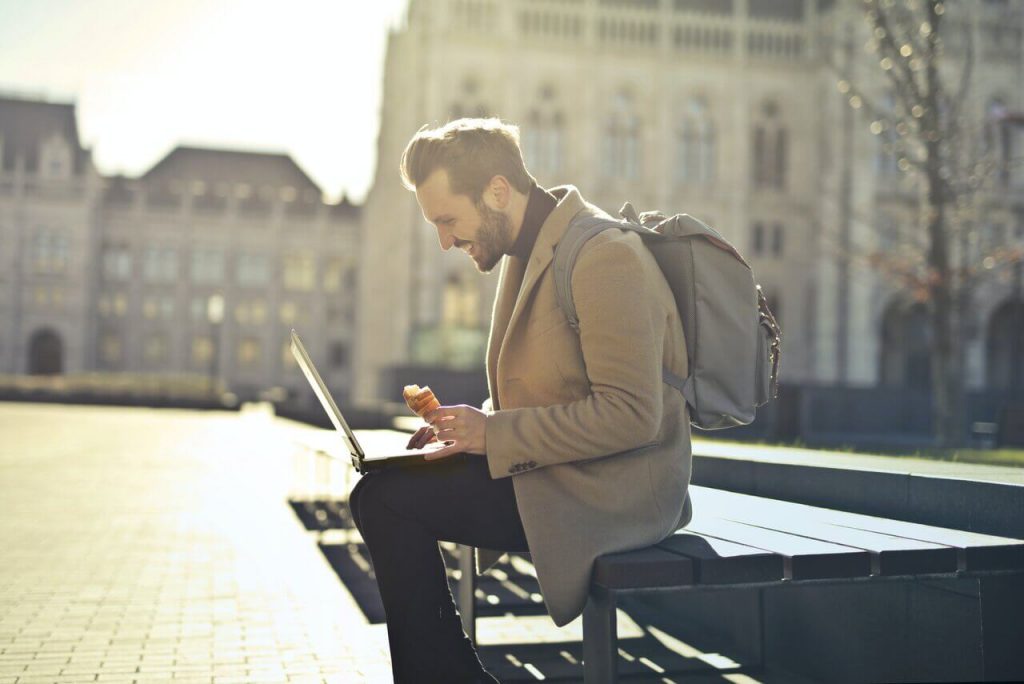 European digital nomads man in brown coat sitting outside on his laptop and eating a croissant