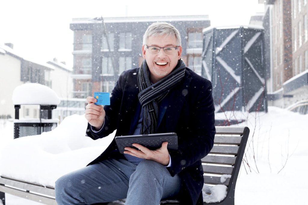 E-resident Jan Lagast holding his blue digital ID card and smiling while sitting on a bench in snowy Tallinn