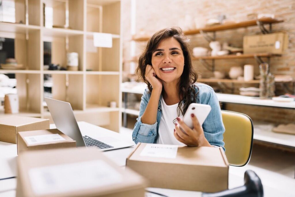 Woman owner of eCommerce micro-enterprise smiling while on her mobile phone and surrounded by delivery packages